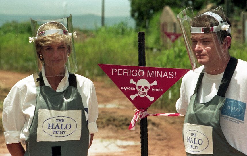 Princess Diana and Paul Heslop standing in Personal Protective Equipment displaying the HALO logo in a minefield in Angola